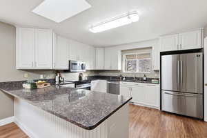 Kitchen with appliances with stainless steel finishes, white cabinets, a skylight, dark stone countertops, and light wood-style floors