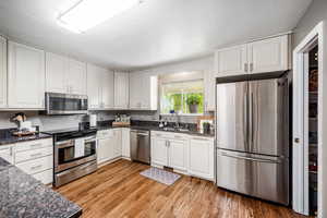 Kitchen with stainless steel appliances, white cabinets, light wood-type flooring, dark stone counters, and a textured ceiling