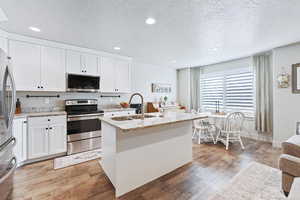 Kitchen with white cabinetry, stainless steel appliances, a textured ceiling, an island with sink, and light stone countertops