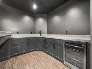 Bar with beverage cooler, gray cabinets, brick floors, and light stone counters