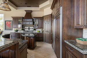 Kitchen featuring tasteful backsplash, glass insert cabinets, dark stone counters, a raised ceiling, and dark brown cabinets