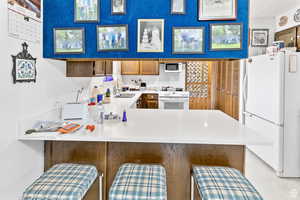 Kitchen with brown cabinetry, white appliances, a breakfast bar area, light countertops, and a peninsula