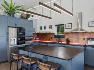 Kitchen featuring appliances with stainless steel finishes, a breakfast bar area, wall chimney range hood, open shelves, and blue cabinetry