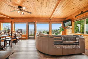 Living room featuring wood-style LVP flooring, a wooden ceiling with exposed beams, wooden walls, a baseboard heating unit, deck access, and a ceiling fan