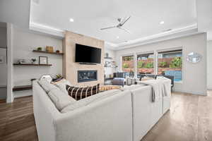 Living area featuring a raised ceiling, ceiling fan, light wood-style flooring, a tiled fireplace, and recessed lighting