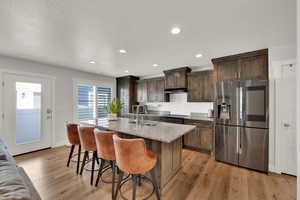 Kitchen featuring stainless steel fridge with ice dispenser, light stone countertops, a center island with sink, recessed lighting, and light wood-style floors
