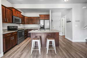Kitchen featuring appliances with stainless steel finishes, a center island with sink, light stone countertops, light wood-type flooring, and dark brown cabinets