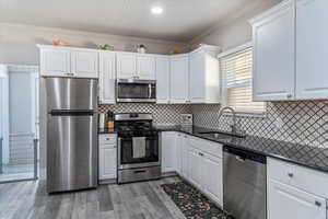Kitchen with appliances with stainless steel finishes, crown molding, dark stone counters, white cabinets, and light wood-type flooring