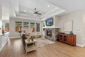 Living area featuring a stone fireplace, a tray ceiling, a ceiling fan, light wood-style floors, and recessed lighting