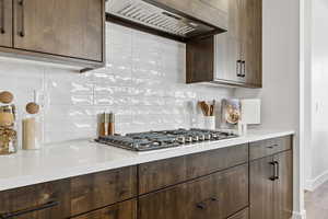 Kitchen with tasteful backsplash, premium range hood, dark brown cabinetry, stainless steel gas cooktop, and light stone counters