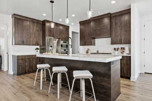 Kitchen with dark brown cabinets, decorative light fixtures, a breakfast bar, a center island with sink, and tasteful backsplash
