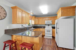 Kitchen featuring white appliances, a peninsula, dark stone counters, a breakfast bar area, and dark wood finished floors