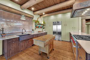 Kitchen with high end appliances, decorative light fixtures, wall chimney range hood, a wooden ceiling with exposed beams, and light wood finished floors