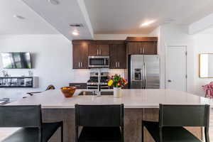 Kitchen with a breakfast bar area, dark brown cabinets, decorative backsplash, stainless steel appliances, and a kitchen island with sink