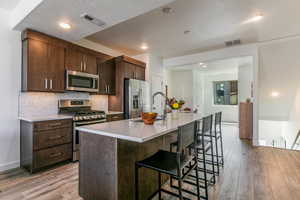 Kitchen with dark brown cabinets, an island with sink, appliances with stainless steel finishes, a kitchen bar, and backsplash