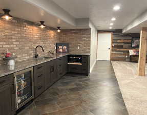 Indoor wet bar featuring wine cooler, light stone countertops, wood walls, stainless steel microwave, and dark brown cabinetry