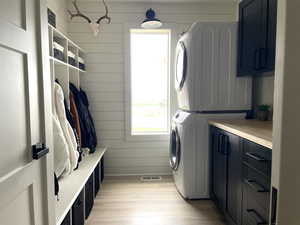 Laundry room featuring wooden walls, plenty of natural light, stacked washing machine and dryer, light wood-style floors, and cabinet space