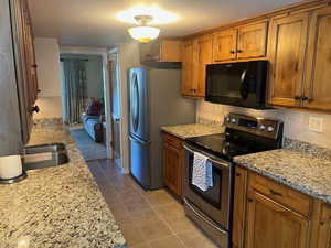 Kitchen with stainless steel appliances, tasteful backsplash, brown cabinetry, light tile patterned flooring, and light stone countertops