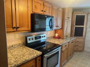 Kitchen featuring stainless steel electric stove, black microwave, light tile patterned floors, backsplash, and brown cabinetry