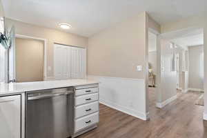 Kitchen with dishwasher, white cabinets, light wood-style floors, wainscoting, and light stone countertops