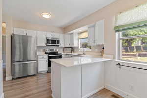 Kitchen with appliances with stainless steel finishes, white cabinetry, a peninsula, light wood-style flooring, and light stone countertops
