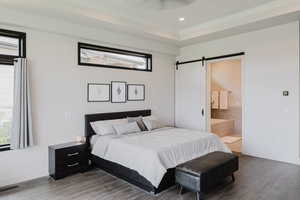 Bedroom featuring a barn door, wood finished floors, ensuite bathroom, recessed lighting, and a raised ceiling