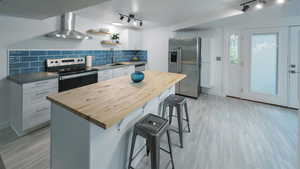 Basement Kitchen with white cabinetry, stainless steel appliances, wall chimney exhaust hood, rail lighting, and a kitchen island