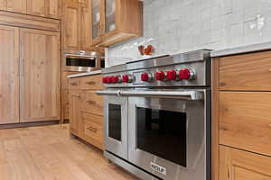 Kitchen with stainless steel appliances, glass insert cabinets, light wood-style flooring, backsplash, and light brown cabinetry
