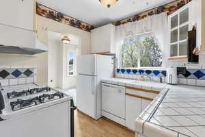 Kitchen featuring tasteful backsplash, white cabinets, white appliances, light wood-type flooring, and tile countertops