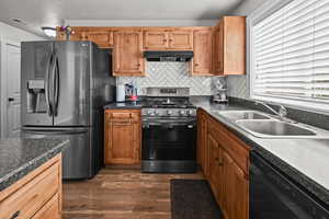 Kitchen with refrigerator with ice dispenser, stainless steel gas range, black dishwasher, backsplash, and dark wood-style floors
