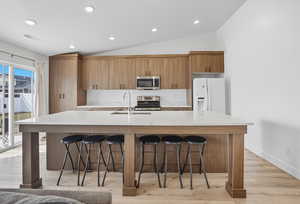 Kitchen featuring brown cabinets, light stone counters, an island with sink, appliances with stainless steel finishes, and vaulted ceiling