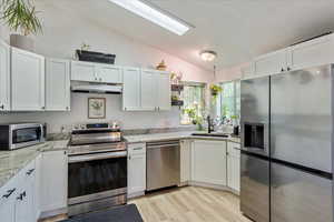 Kitchen with appliances with stainless steel finishes, vaulted ceiling, white cabinets, light wood-style floors, and light stone counters