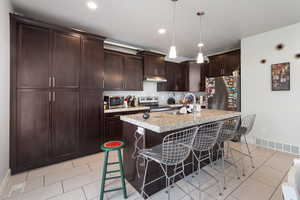 Kitchen featuring a kitchen breakfast bar, a kitchen island with sink, dark brown cabinetry, decorative light fixtures, and light tile patterned flooring