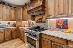 Kitchen featuring high end stove, light stone counters, premium range hood, brown cabinets, and decorative backsplash