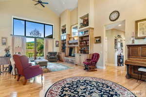 Sitting room with high vaulted ceiling, light wood finished floors, ceiling fan, and a fireplace with raised hearth