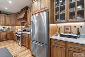 Kitchen featuring stainless steel appliances, brown cabinets, glass insert cabinets, custom range hood, and light wood-type flooring