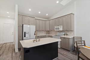 Kitchen with white appliances, gray cabinetry, light wood-style flooring, lofted ceiling, and a kitchen island with sink
