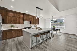 Kitchen with hanging light fixtures, a breakfast bar area, an island with sink, backsplash, and light stone countertops