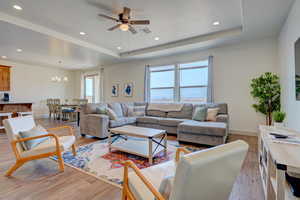 Living room featuring light wood-type flooring, recessed lighting, a ceiling fan, a raised ceiling, and a chandelier