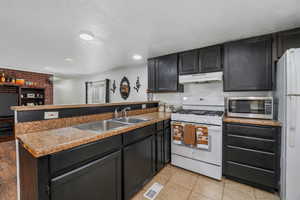 Kitchen featuring dark cabinetry, white appliances, a peninsula, a textured ceiling, and under cabinet range hood
