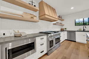 Kitchen with open shelves, appliances with stainless steel finishes, premium range hood, light wood-style floors, and decorative backsplash