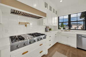 Kitchen with custom exhaust hood, stainless steel appliances, white cabinetry, light wood-style floors, and glass insert cabinets