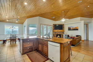 Kitchen featuring a fireplace, a center island with sink, light stone counters, dishwasher, and hanging light fixtures