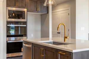 Kitchen featuring dark brown cabinetry, appliances with stainless steel finishes, light stone counters, and decorative light fixtures