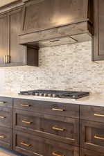 Kitchen with premium range hood, dark brown cabinetry, light stone countertops, stainless steel gas cooktop, and decorative backsplash