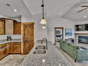 Kitchen with vaulted ceiling, pendant lighting, brown cabinetry, light stone counters, and a glass covered fireplace