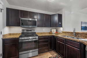 Kitchen with stainless steel appliances, dark brown cabinetry, light stone counters, a textured ceiling, and light wood-style floors