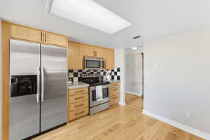 Kitchen featuring light brown cabinetry, stainless steel appliances, decorative backsplash, and light wood flooring