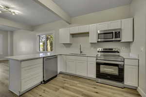 Kitchen with appliances with stainless steel finishes, white cabinets, beam ceiling, light wood-style flooring, and a peninsula