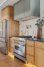 Kitchen with high end appliances, light wood-style flooring, custom exhaust hood, tasteful backsplash, and light brown cabinetry